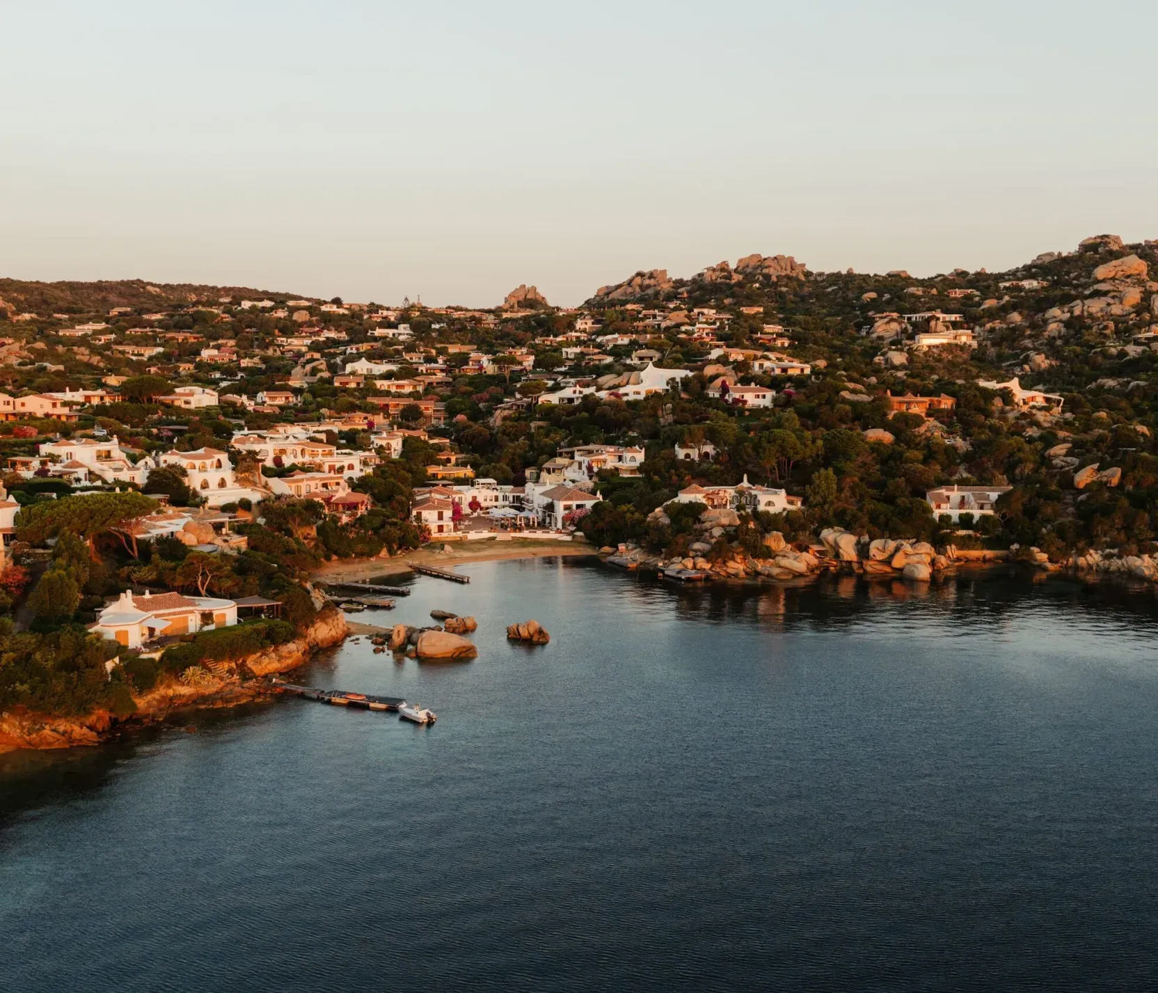 A coastal village with white houses nestled among rocky hills, seen at sunset. The tranquil sea reflects the golden light, creating a serene atmosphere.