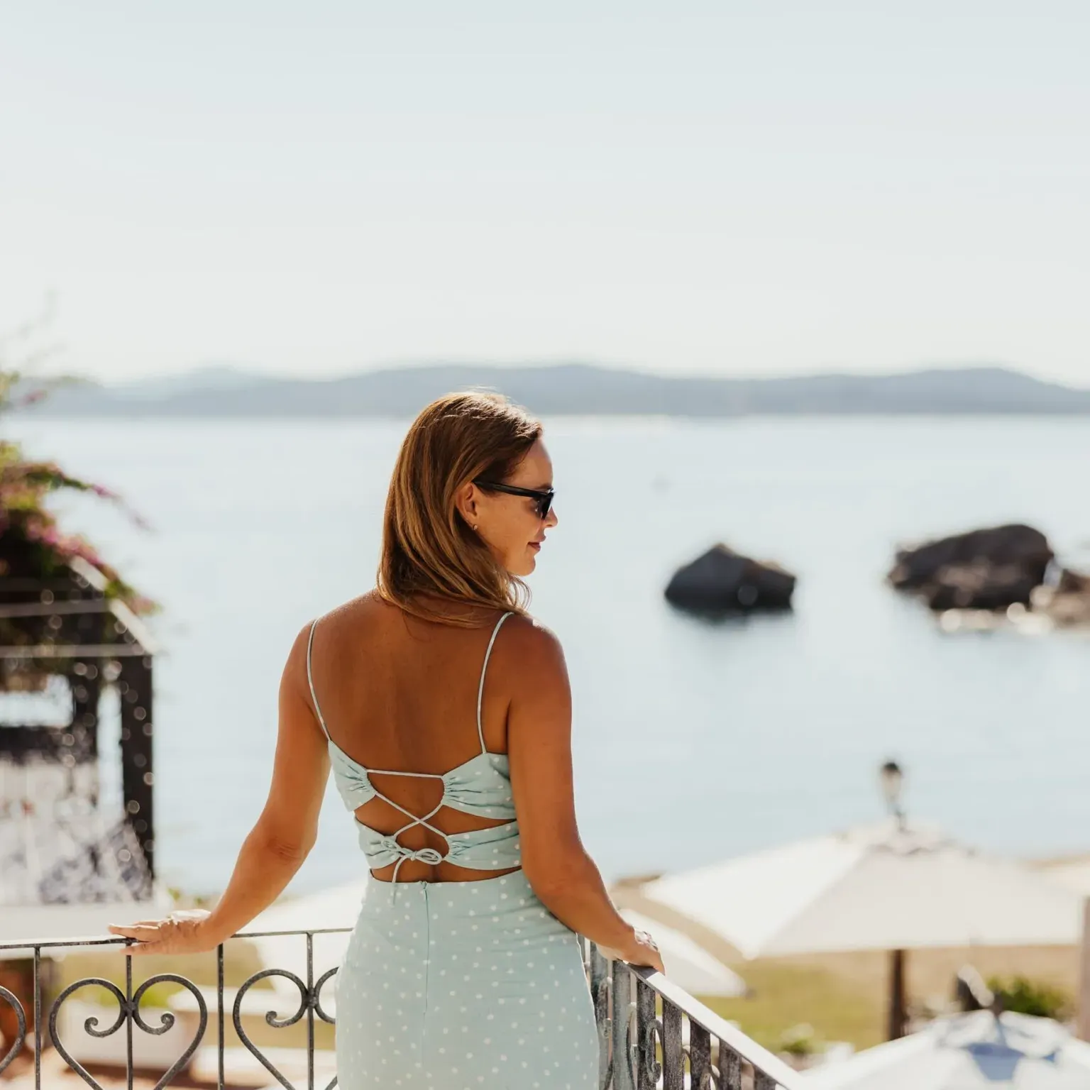 A woman in a light blue dress stands on a balcony overlooking a serene beach with gentle waves and distant mountains, exuding a relaxed summer vibe.