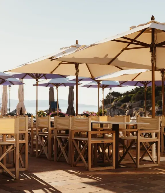 Sunny outdoor café with wooden tables and chairs under large beige umbrellas. Vibrant purple flowers, ocean view, and clear sky create a relaxed ambiance.