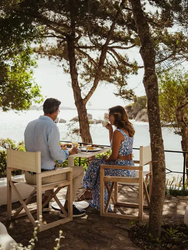 A couple enjoys a peaceful outdoor meal with a stunning seaside view in Porto Rafael, surrounded by lush greenery and warm sunlight filtering through the trees.