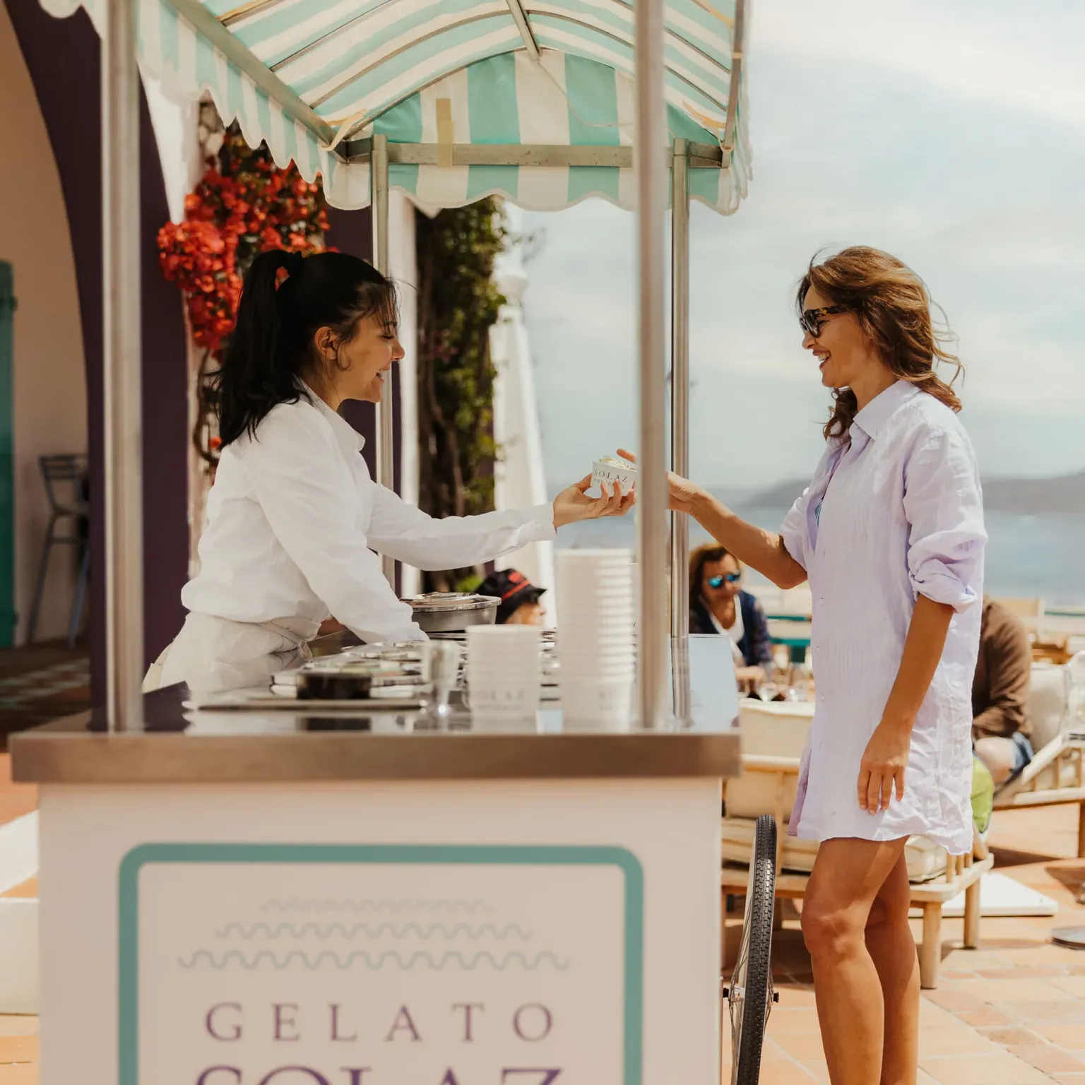 A woman in a light dress buys gelato from a vendor under a striped canopy at a seaside location. Both are smiling, creating a cheerful atmosphere.