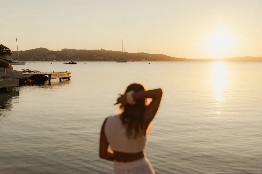 A woman in white stands on a calm beach at sunset, gazing at the horizon. Boats float on tranquil water, creating a serene and peaceful atmosphere.