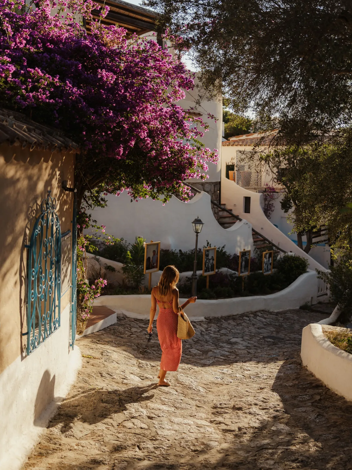 A woman in a red dress walks down a sunlit cobblestone path, surrounded by vibrant purple flowers and white stucco buildings, conveying a serene, warm atmosphere in Porto Rafael.