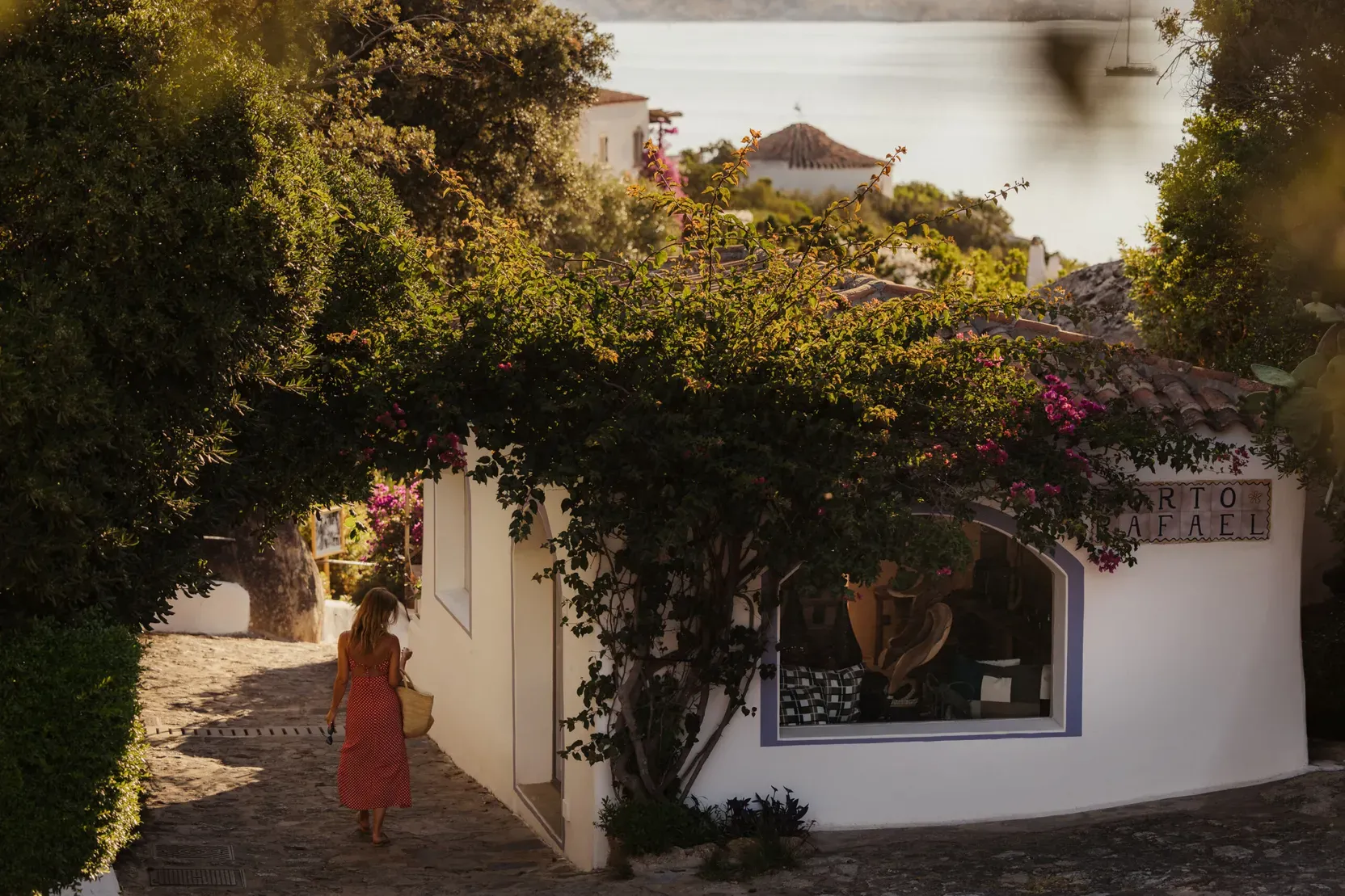 A woman in a red dress walks down a quaint cobblestone street lined with lush greenery and flowers. White buildings and a glimpse of the sea enhance the serene, Mediterranean atmosphere.
