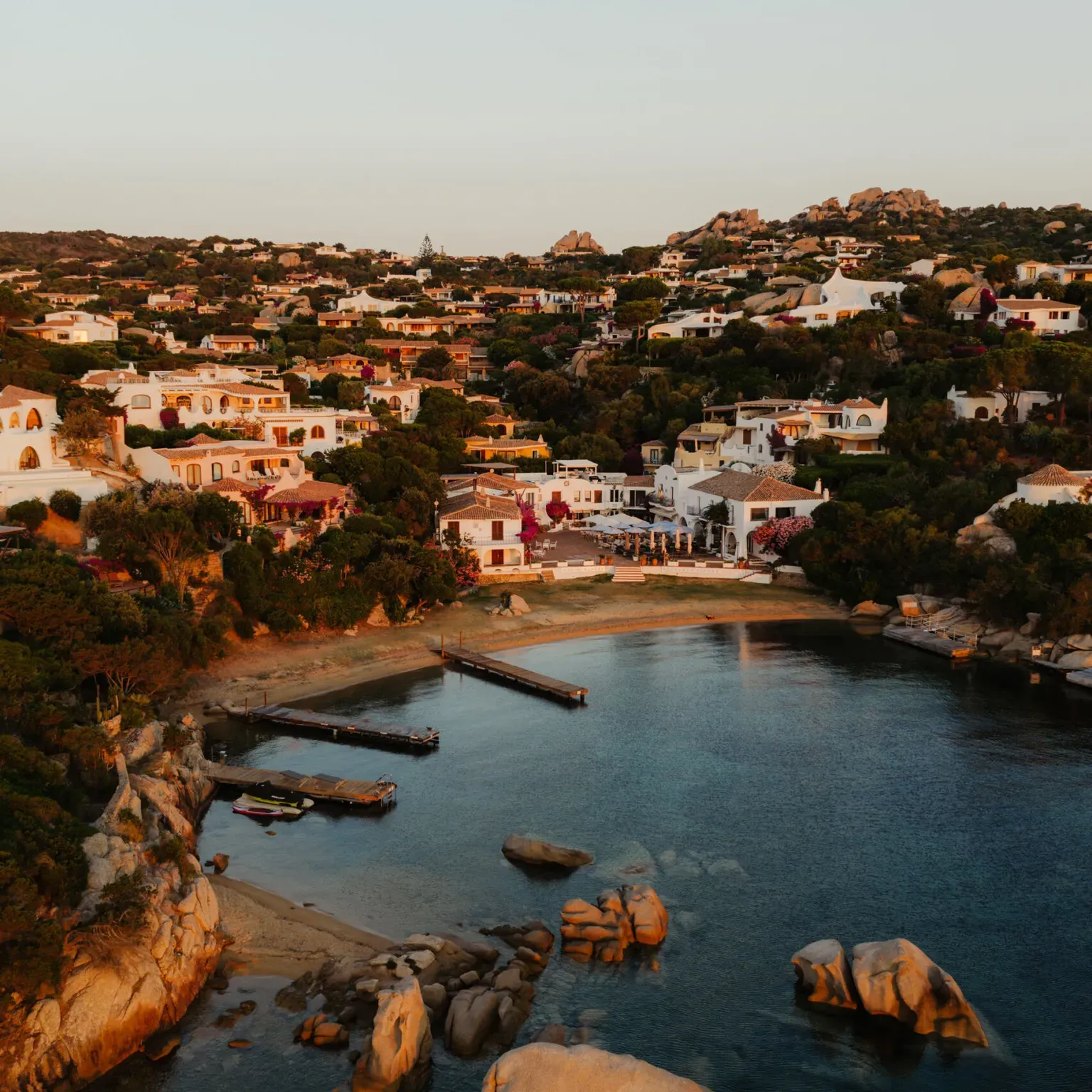 Coastal village at sunset with white Mediterranean-style houses on a hillside, surrounded by lush greenery.