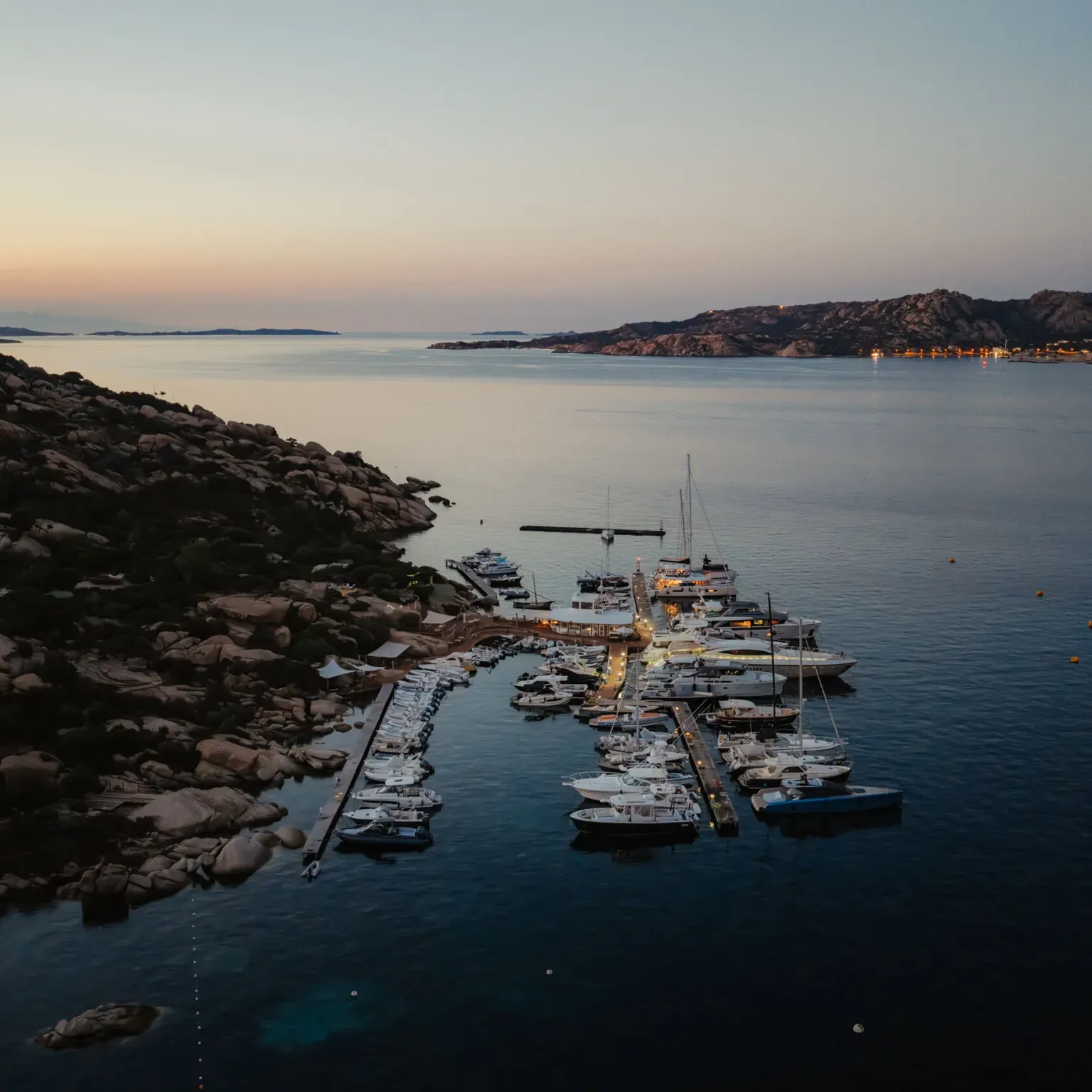 A serene marina at dusk, with yachts docked alongside rocky shores. The calming sea reflects the fading light of the sunset, creating a tranquil ambiance.