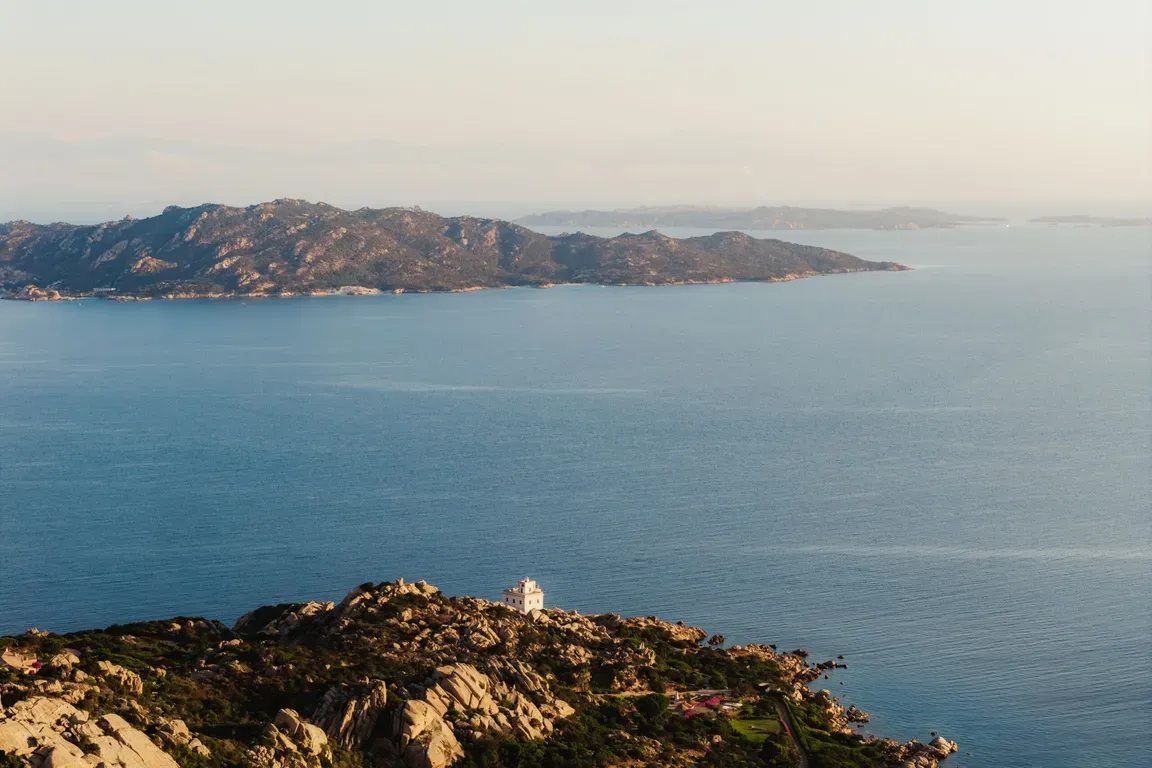 A serene coastal landscape with a rocky foreground and a small white lighthouse, overlooking a calm blue sea. Distant mountains provide a tranquil backdrop.