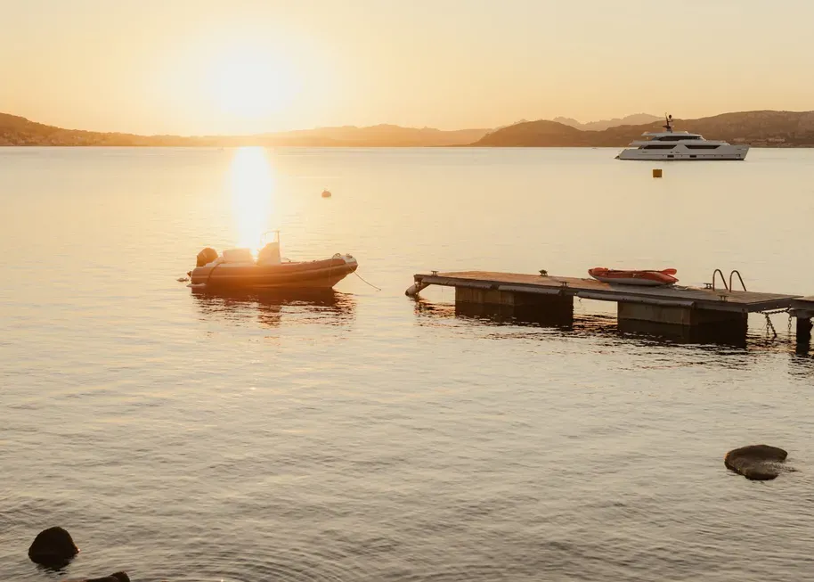 Serene sunset over calm water with a small boat near a wooden dock in the foreground. A yacht is visible in the distance. Tranquil ambiance.