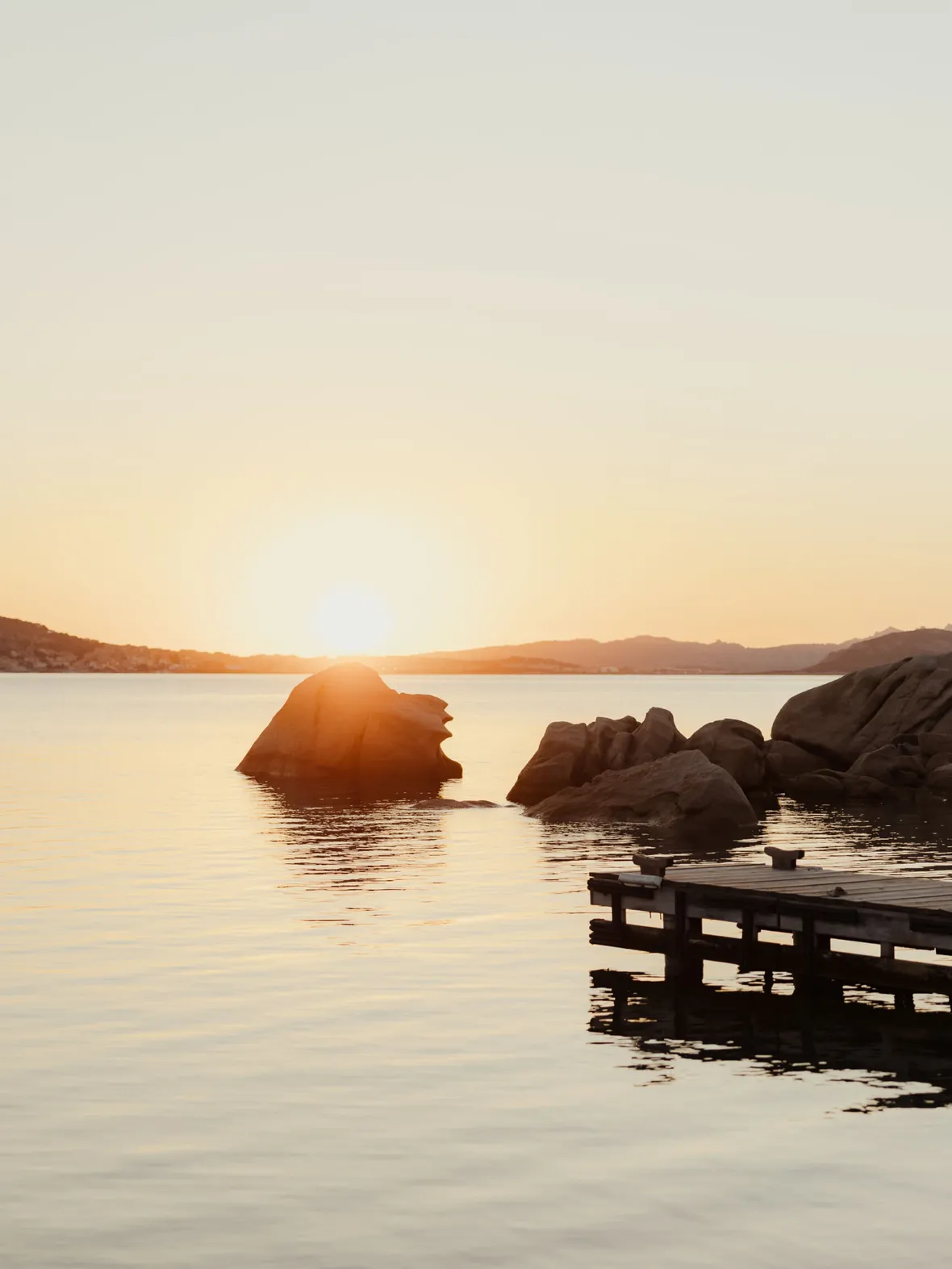 Sunset over calm water with silhouetted rocks and a wooden pier. The sky is a soft gradient of orange, creating a peaceful and serene atmosphere.