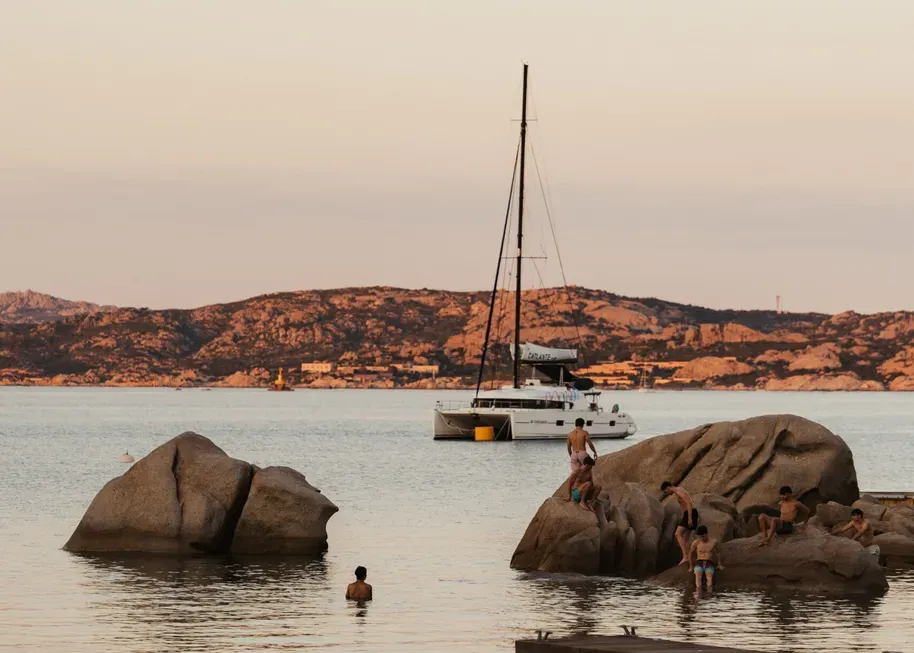 Calm beach scene at sunset with people on rocks and swimming. A sailboat floats in the distance against a backdrop of hills, creating a serene atmosphere.