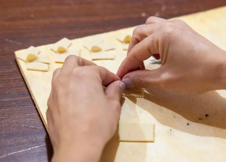 Hands are delicately folding small pasta shapes on a wooden board. The setting conveys a sense of careful craftsmanship and focus.