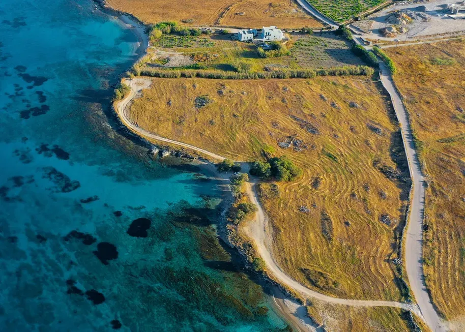 Aerial view of a coastal landscape with turquoise waters and rocky shores. Golden brown fields and small buildings line the coast, creating a serene, picturesque scene.