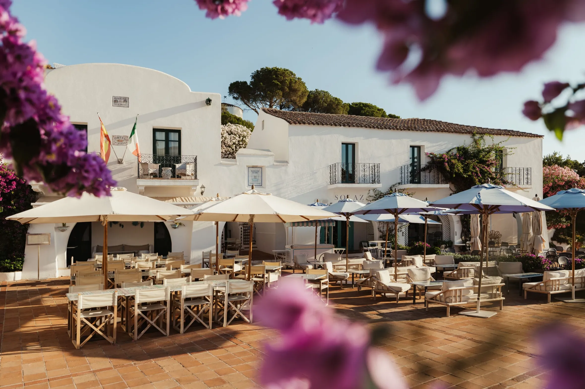 Charming courtyard of a white Mediterranean villa with shaded outdoor dining tables. Purple flowers frame the view. The atmosphere is tranquil and inviting.