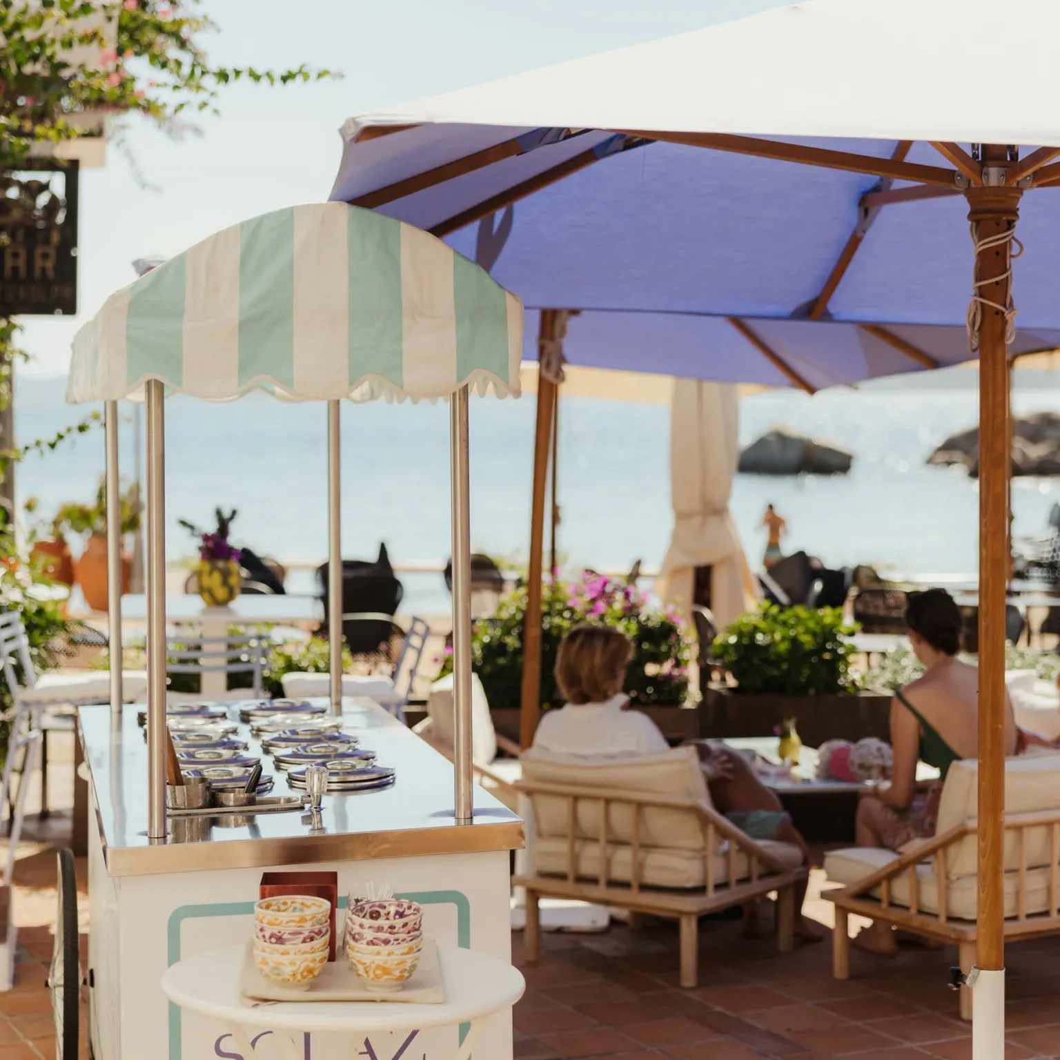 Beachside café setting with people relaxing under large blue umbrellas. Ice cream cart with colorful cups in foreground, ocean view in background. Calm and inviting atmosphere.