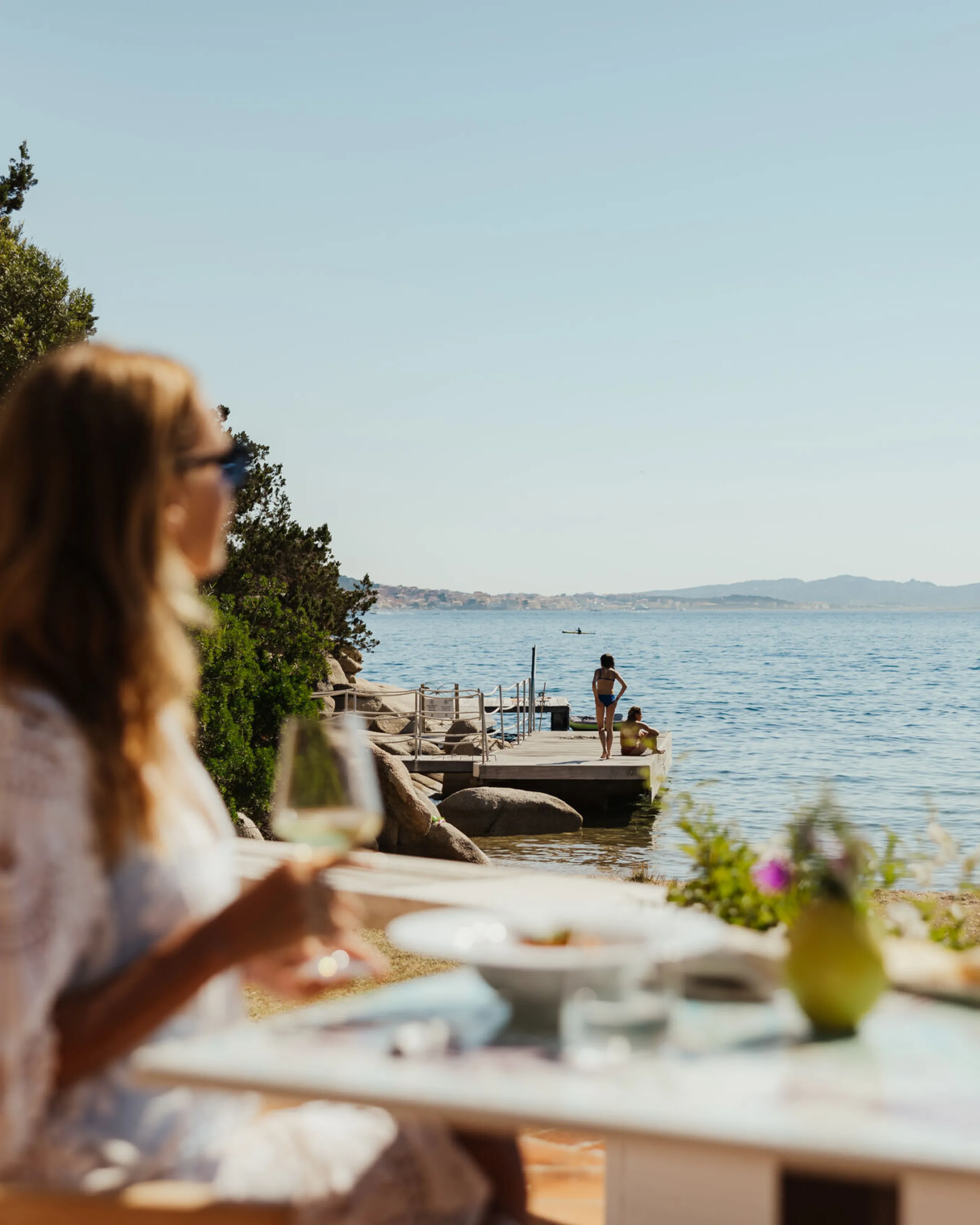 A woman in sunglasses enjoys a seaside meal at a table under clear skies. The scene is tranquil, featuring distant hills and calm water.